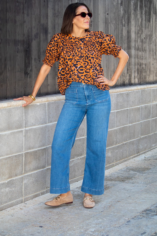 Woman wearing a leopard print top and blue jeans standing against a gray wall.