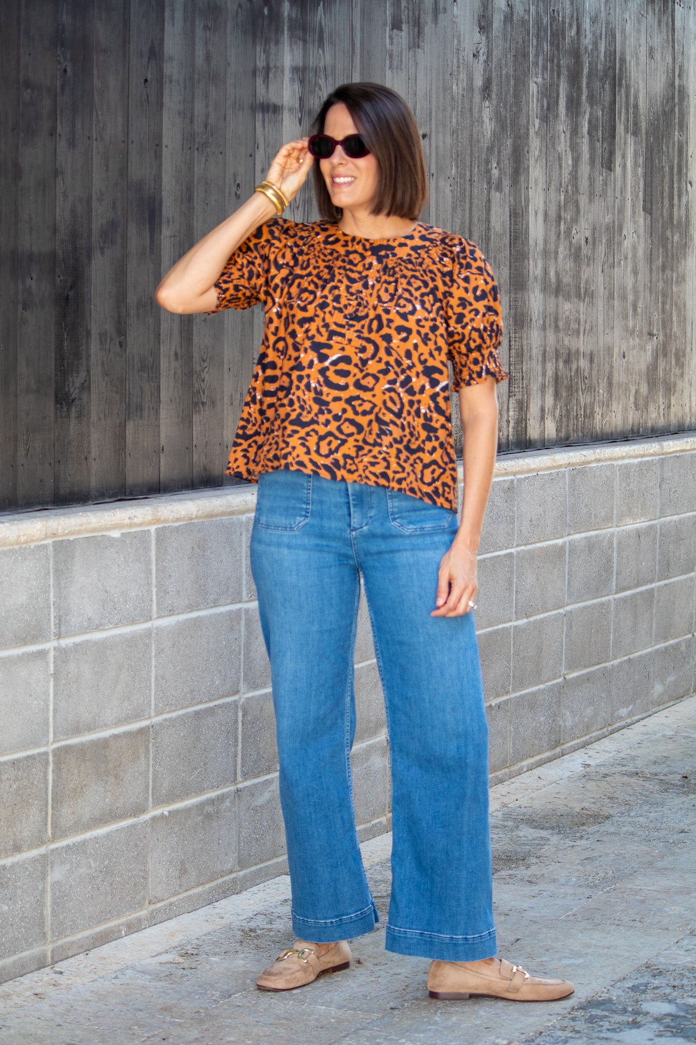 Woman wearing a leopard print top and blue jeans standing against a gray wall.