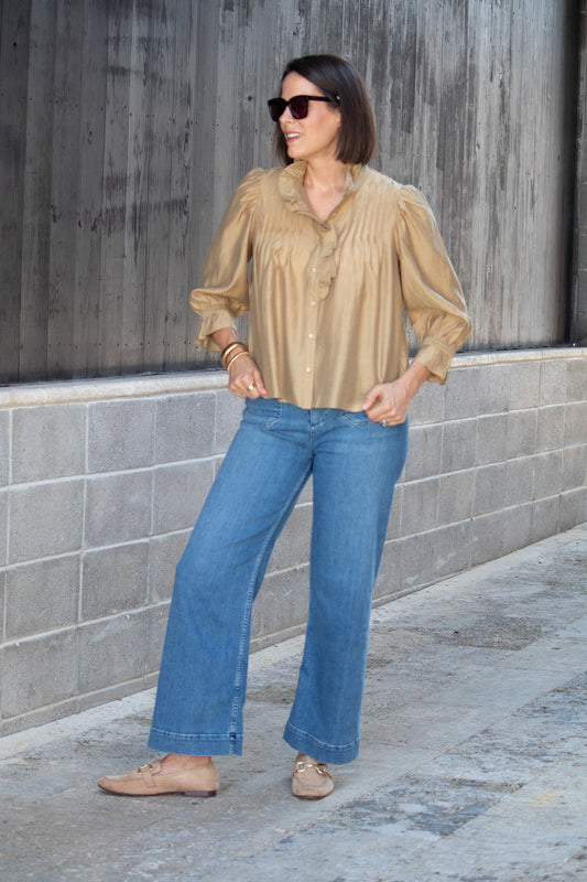 Woman wearing a neutral blouse and blue jeans standing against a gray wall.