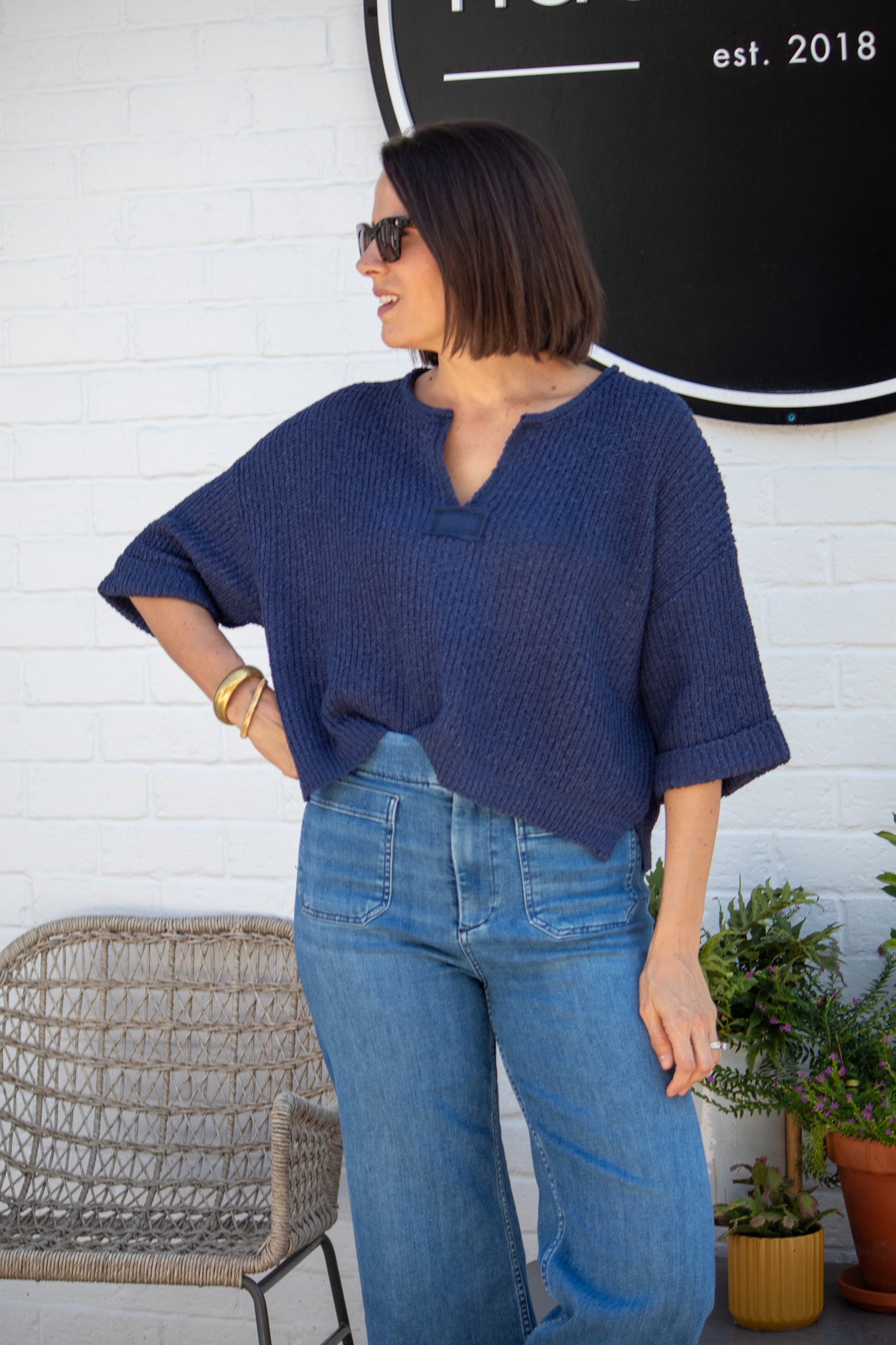 Woman wearing a blue sweater and jeans standing in front of a white wall with a circular sign.