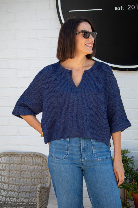 Woman wearing a blue textured top and jeans standing against a white brick wall.