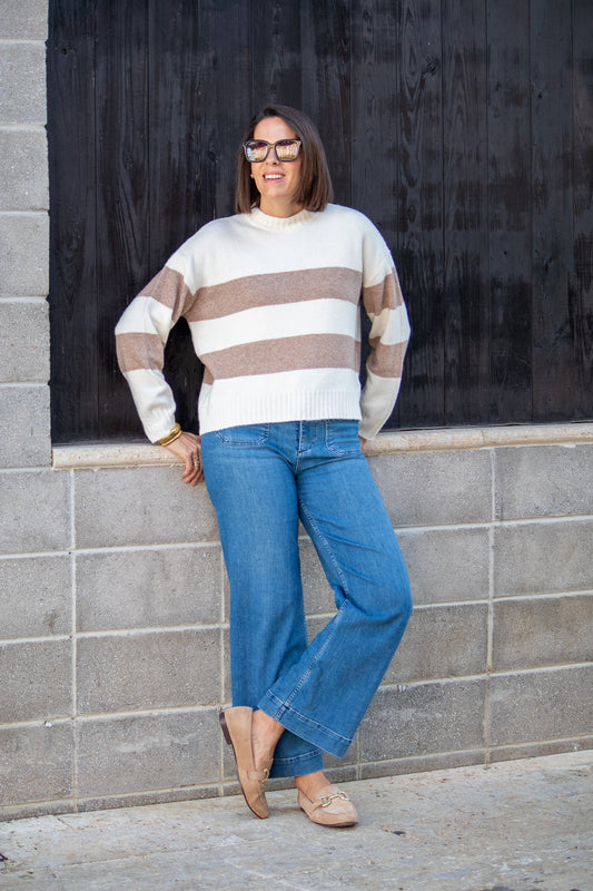 Person wearing a striped sweater and blue jeans leaning against a gray wall.