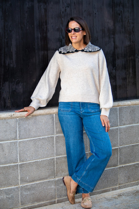 Woman wearing a tan  sweater with a decorative collar and blue jeans against a black wall.