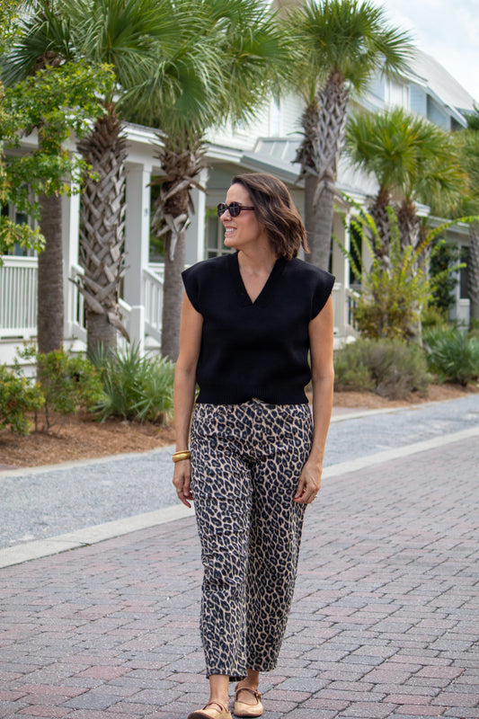 Woman wearing a black top and leopard print pants standing on a brick path with palm trees in the background.