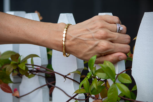 Hand with gold bracelet and ring on a white picket fence with greenery