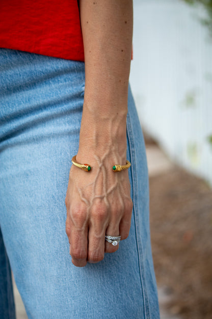 Close-up of a person's wrist wearing a gold bracelet with green stones and a ring, against a blurred natural background.