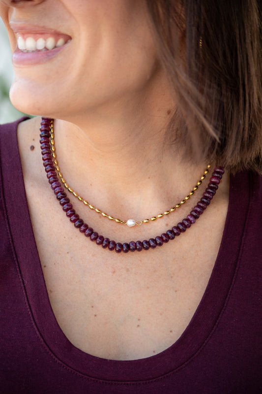 Close-up of a woman wearing a gold necklace and a berry color beaded necklace.