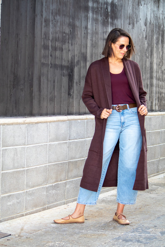 Woman wearing a long brown cardigan, burgundy top, and light blue jeans against a gray wall.