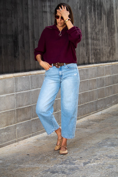 Woman wearing a burgundy sweater and light blue jeans against a gray wall.