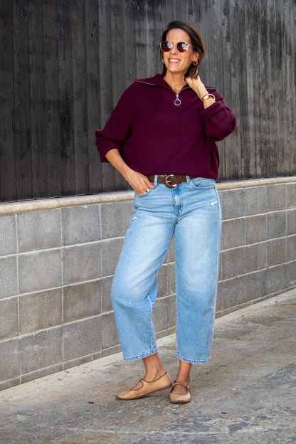 Woman wearing a purple sweater and light blue jeans standing against a gray wall.