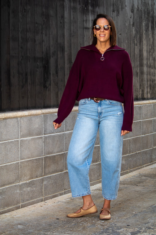 Woman wearing a maroon sweater and light blue jeans standing against a gray wall.