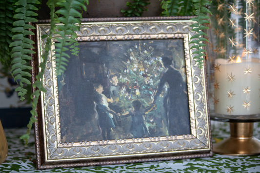Framed artwork of a group of people in a gathered around a Christmas Tree on a decorative table.