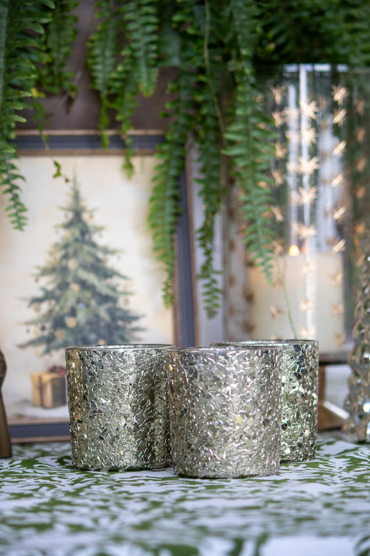 Silver textured candles on a decorative table with plants and Christmas decorations in the background.