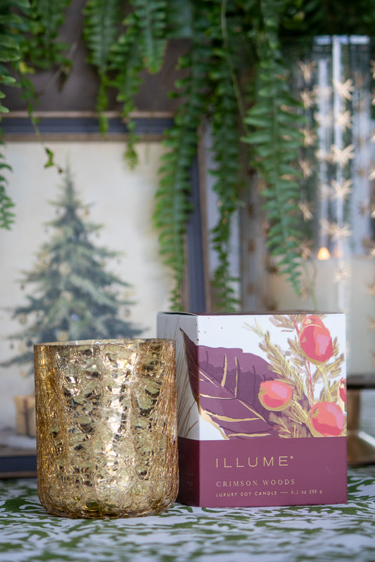 Gold candle and 'Illume' candle packaging on a decorative surface with plants and lights in the background.