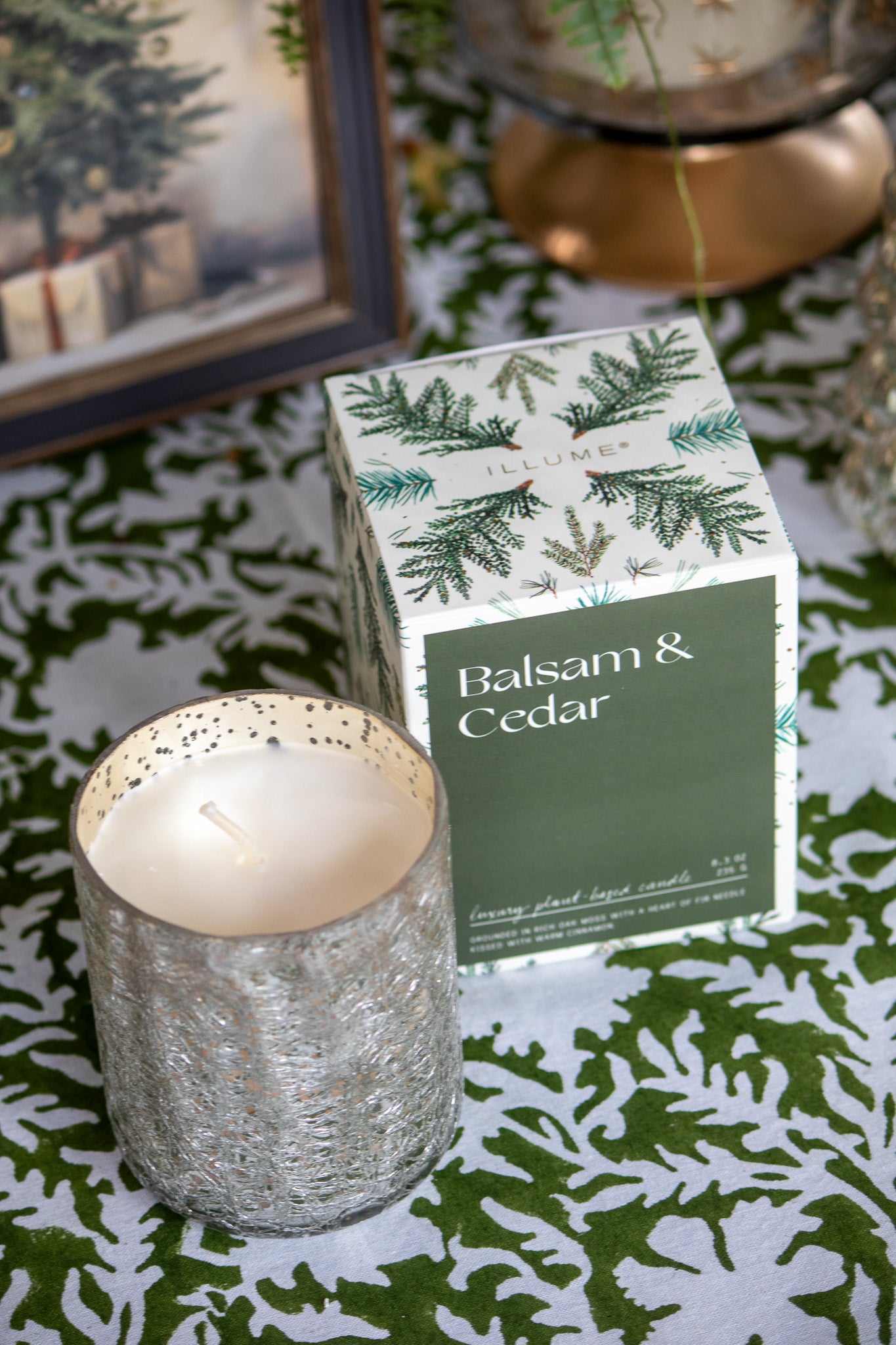 Candle in a textured holder next to a box labeled 'Balsam & Cedar' on a decorative surface.