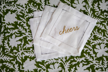 Set of white cocktail napkins with 'cheers' embroidery on a green floral patterned background