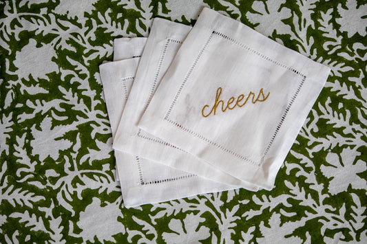 Set of white cocktail napkins with 'cheers' embroidery on a green floral patterned background