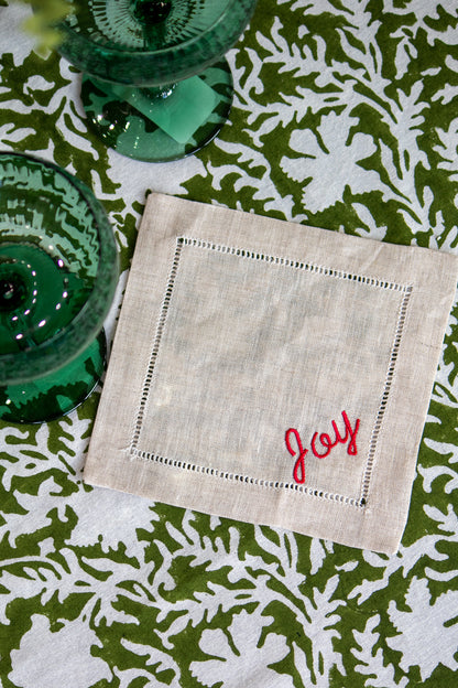 Linen coasters with 'Joy' embroidery on a green and white patterned tablecloth.