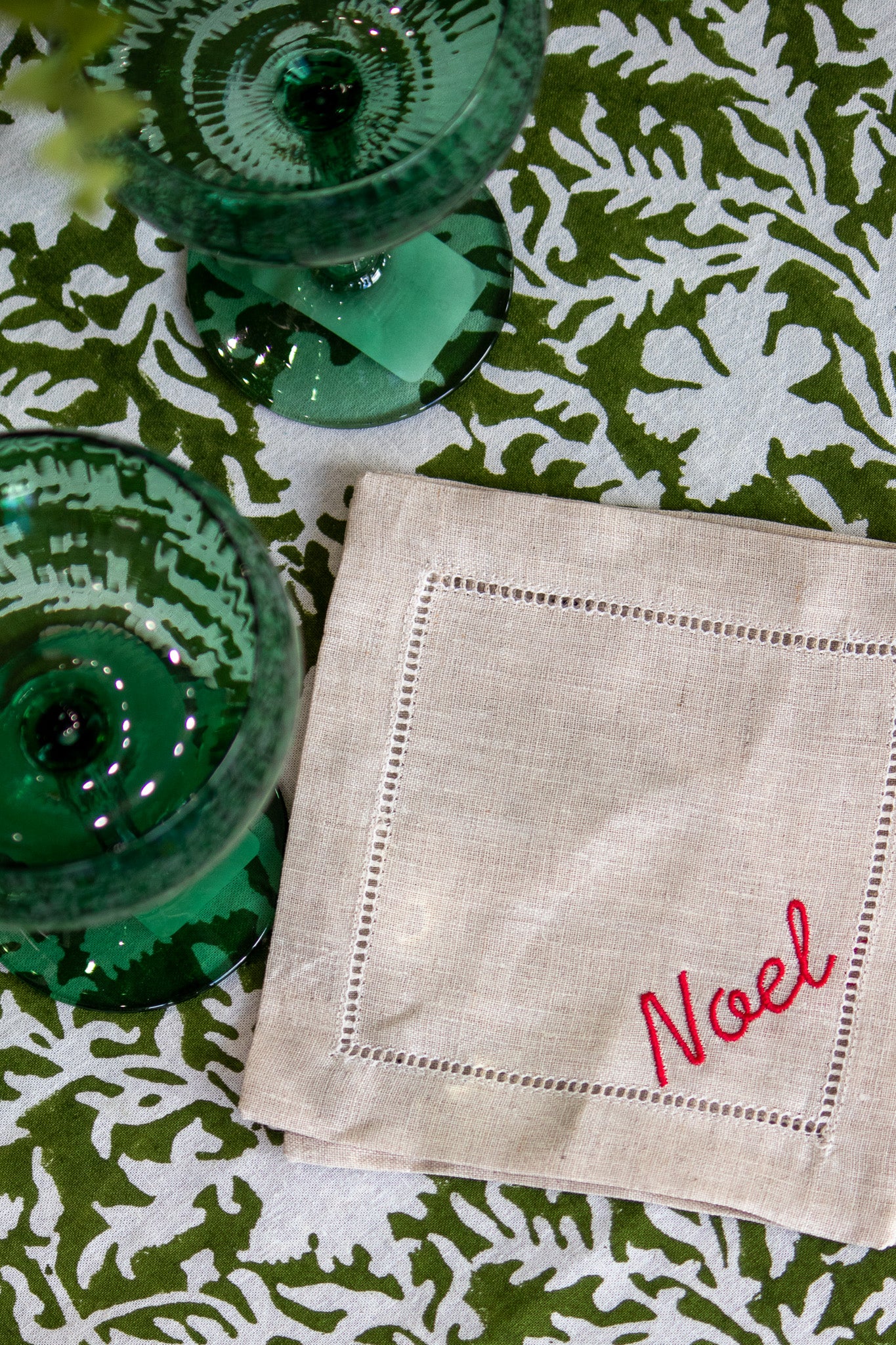 Beige linen coaster with 'Noel' embroidery on a green patterned tablecloth.