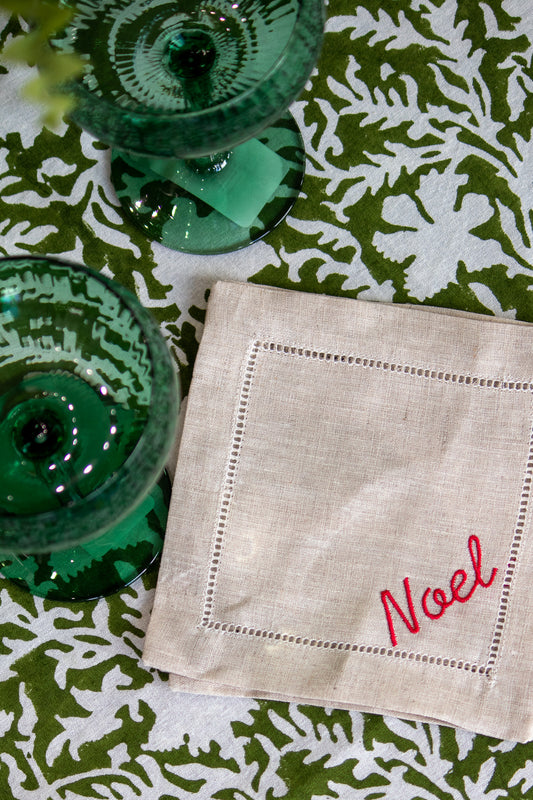 Beige linen coaster with 'Noel' embroidery on a green patterned tablecloth.