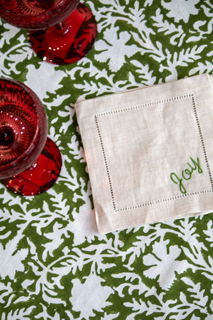 Coaster with 'Joy' embroidery on a green floral tablecloth with red glassware.