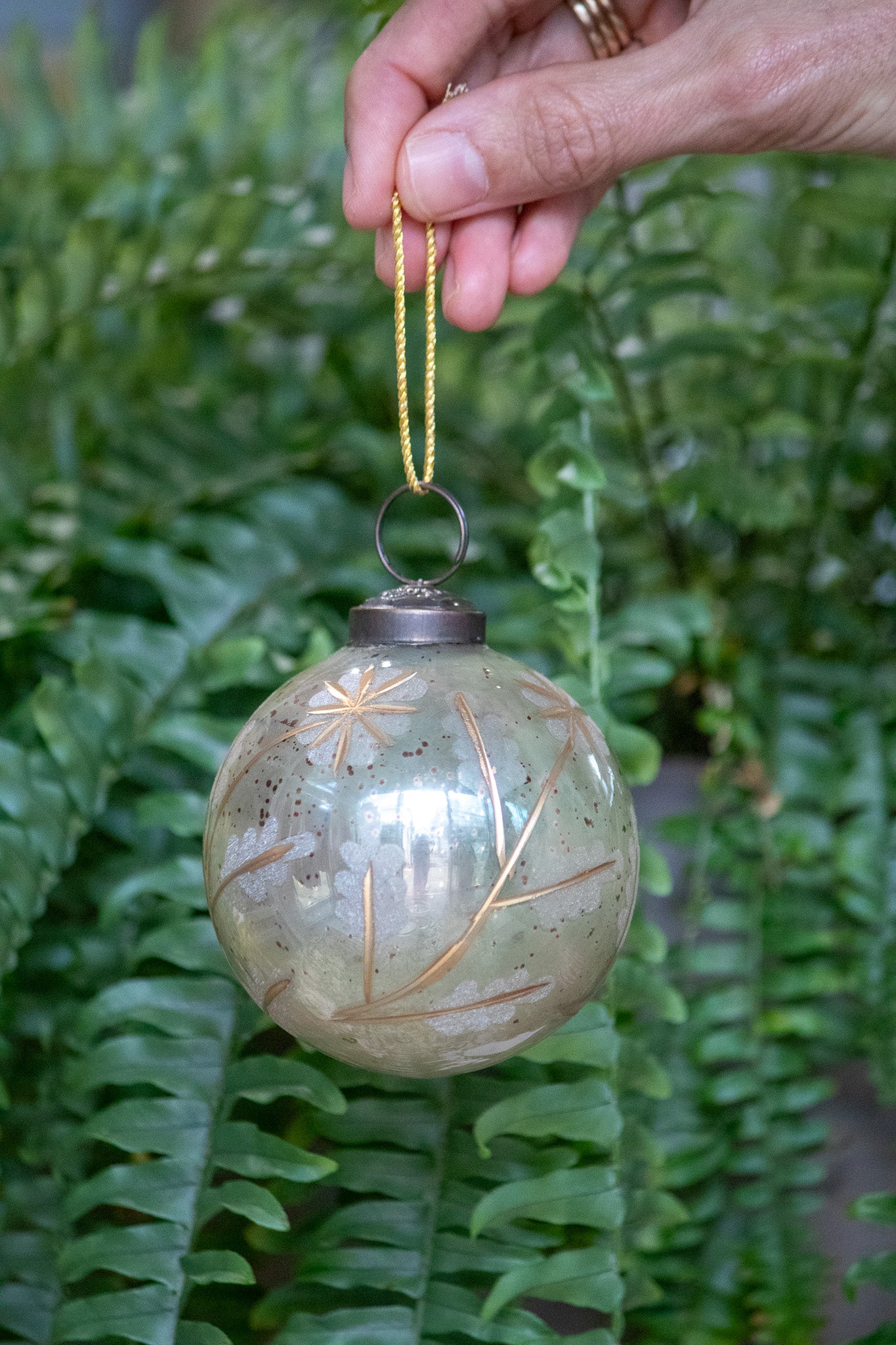 Decorative glass ornament with gold leaf patterns held by a hand against a green fern background