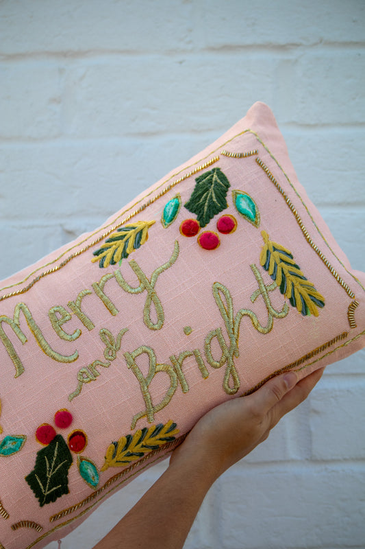 Pink pillow with 'Merry and Bright' embroidery and festive decorations against a light background