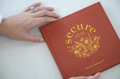 Red book titled 'secure' with yellow text and floral design on a white surface, held by a hand.