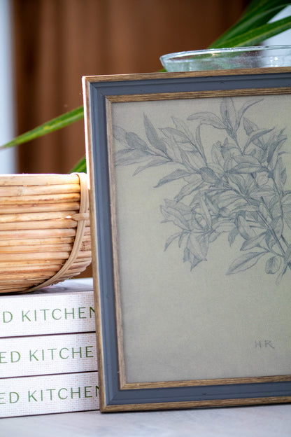 Framed botanical print next to a stack of books on a surface with a plant in the background.