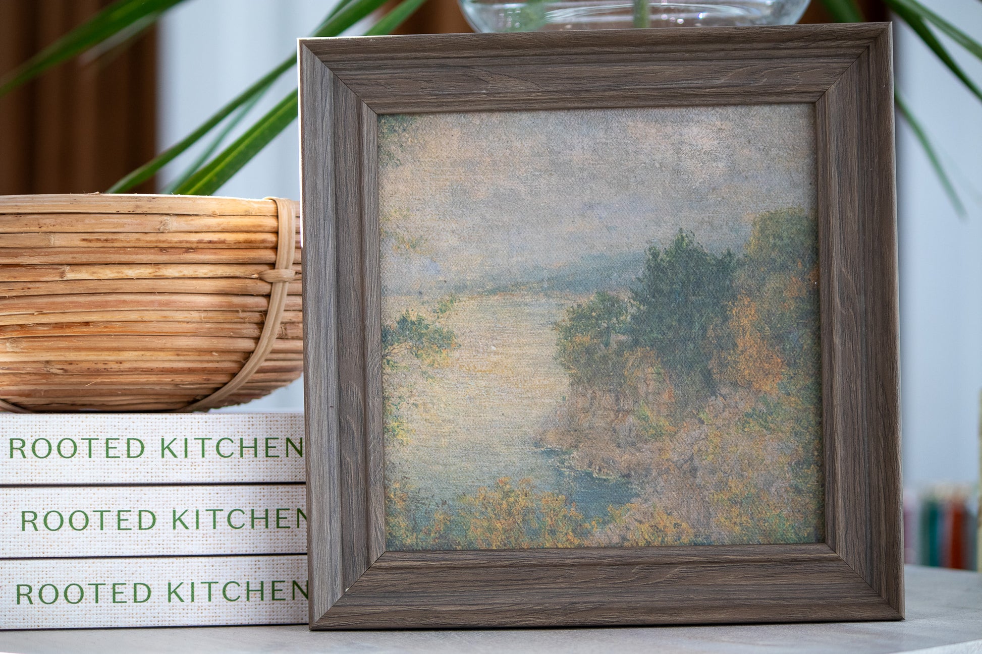 Framed landscape painting on a surface with 'Rooted Kitchen' books in the background
