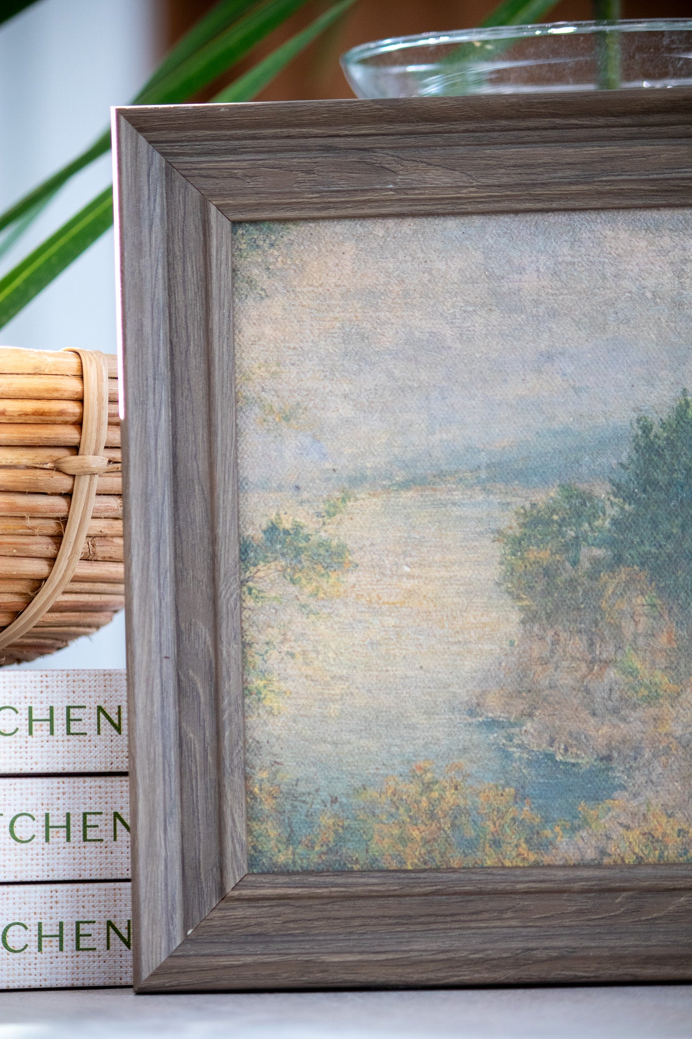 Framed landscape painting on a surface with a basket and kitchen-themed box in the background.
