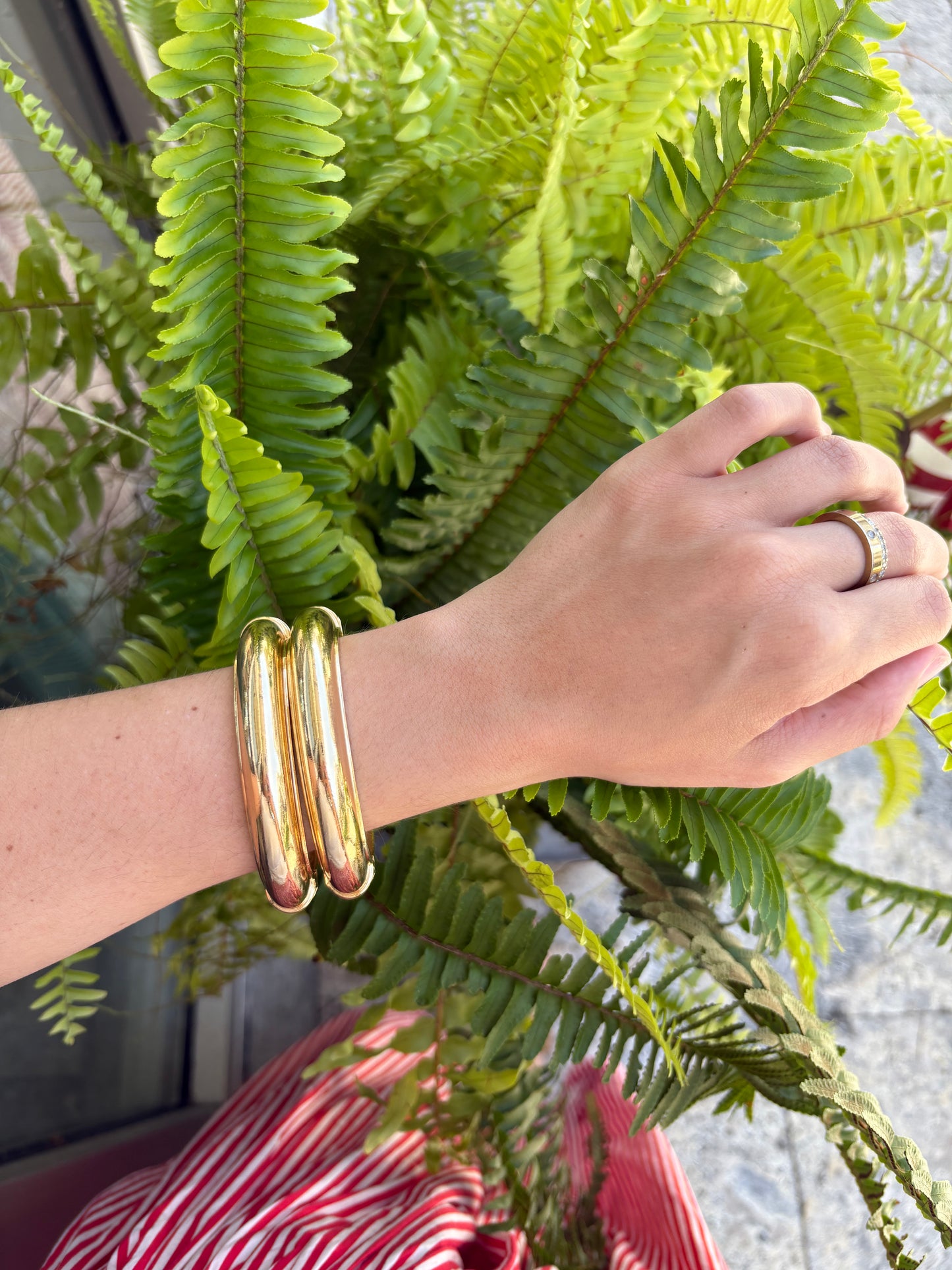 A close-up of a person's wrist wearing three polished gold bangles, with green fern leaves in the background and a red-and-white striped garment visible below.