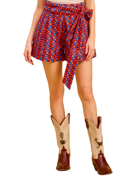 A woman stands against a warm white studio backdrop wearing a cream crochet halter top, a red and blue patterned wrap skirt, and brown cowboy boots with white stitching.