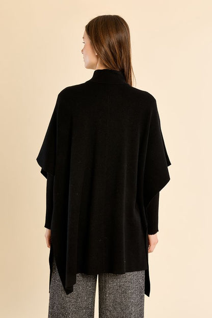 A woman shown from the back wearing a black draped poncho-style top and gray textured pants, standing against a plain beige studio background.