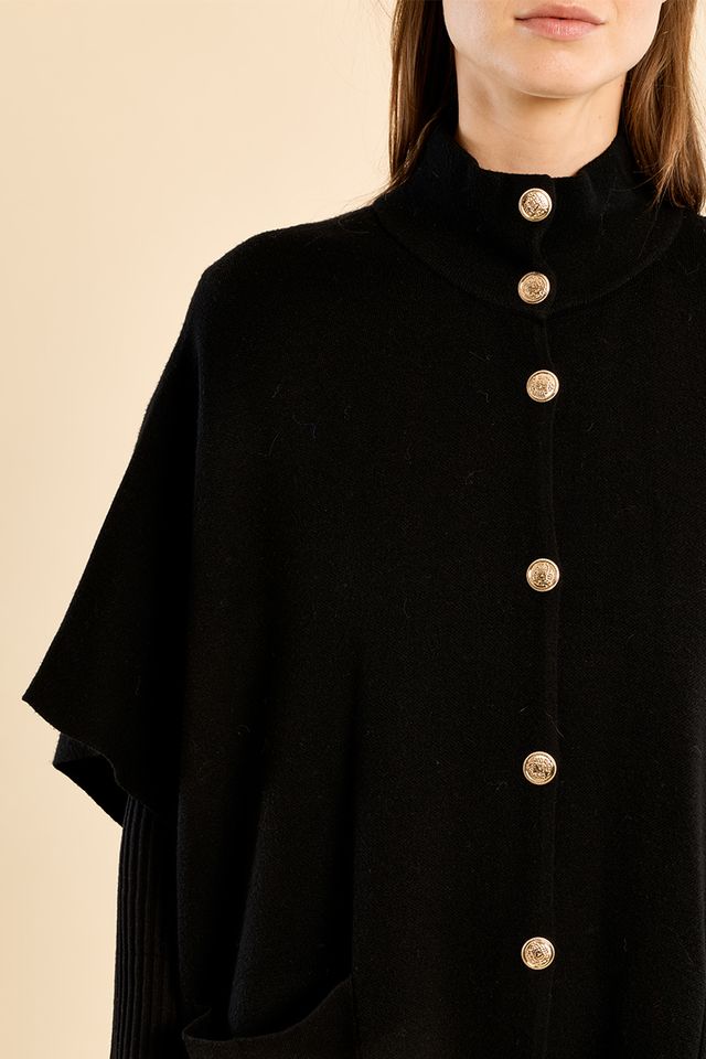 A close-up of a model wearing a black high-neck cape-style top with large gold-tone buttons down the front, set against a solid beige background.