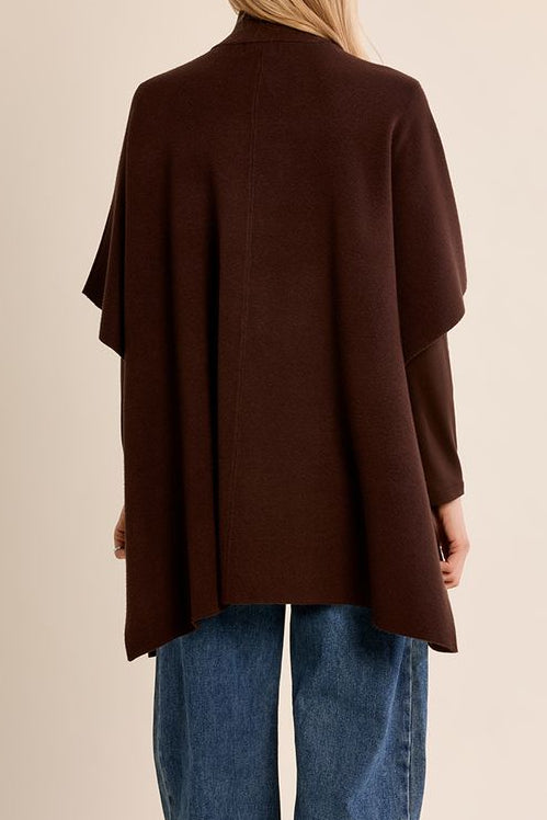 A woman viewed from behind wearing a dark brown open-front poncho-style cardigan over blue jeans, standing against a plain beige studio background.