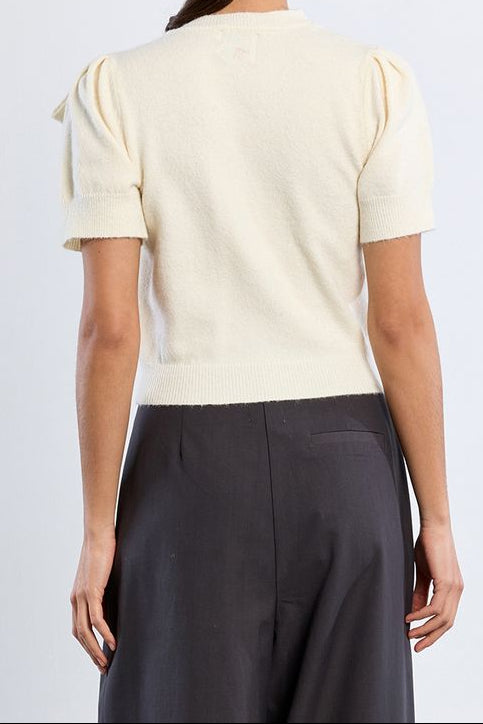 A woman shown from the back wearing a cream-colored short-sleeve knit top with puffed shoulders and dark charcoal wide-leg trousers, set against a plain light gray studio background.