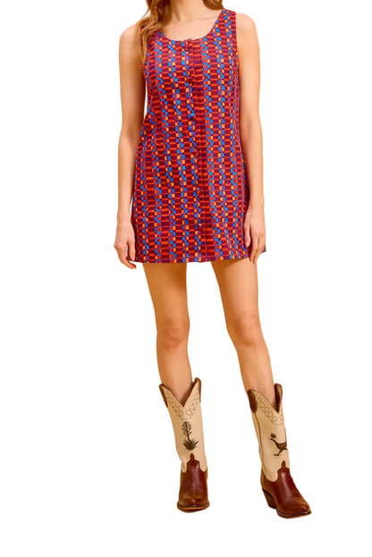 A woman stands against a warm white studio backdrop wearing a sleeveless red and blue patterned mini dress with brown and cream cowboy boots.