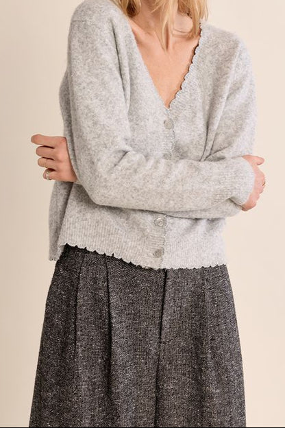 A woman wearing a light gray heathered knit cardigan with a scalloped V-neck and button front, paired with a dark gray textured skirt, against a solid beige background.