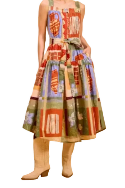 A woman modeling a sleeveless midi dress with a colorful patchwork print in red, green, blue, and beige tones, paired with beige ankle boots, against a solid warm beige studio background.