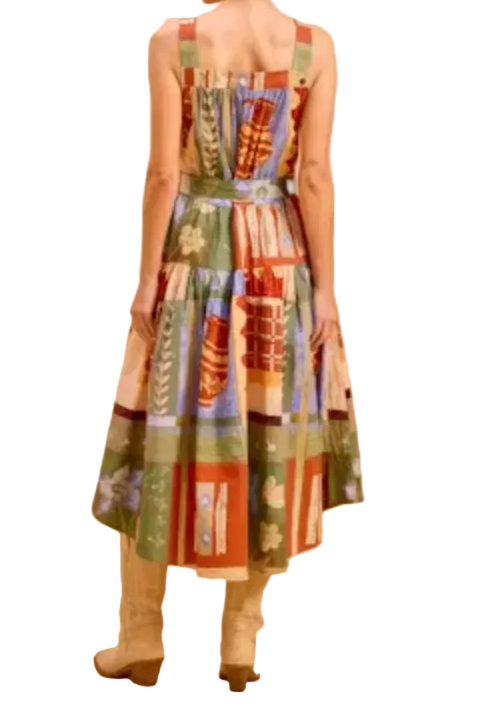 A woman stands with her back to the camera wearing a sleeveless midi dress with a colorful patchwork print in green, red, orange, and blue, paired with beige boots, against a solid warm beige studio background.