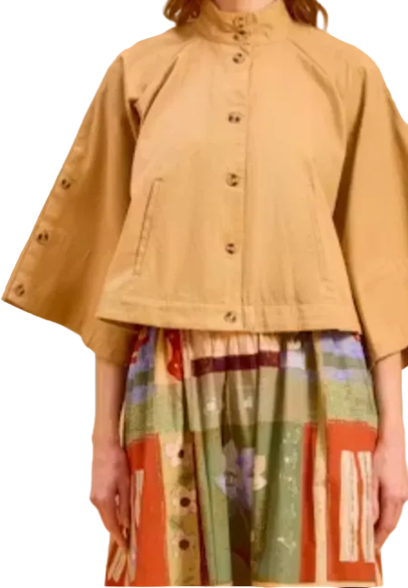 A woman modeling a tan cropped jacket with wide sleeves and decorative snap details on the sleeves, paired with a colorful patterned skirt, against a solid tan background.