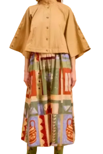 A model stands against a solid beige studio backdrop wearing a tan cropped cape jacket with wide sleeves, a long multicolor patterned skirt, and green boots.