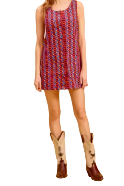 A woman modeling a sleeveless, scoop-neck mini dress with a red, blue, and white geometric pattern, paired with brown and white cowboy boots, set against a solid beige studio backdrop.