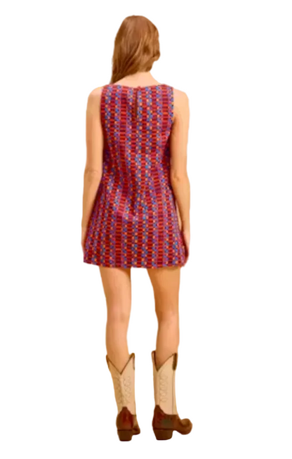 A woman viewed from behind wearing a sleeveless, knee-length dress with a red, blue, and white geometric pattern, paired with brown and cream cowboy boots, against a solid beige studio backdrop.