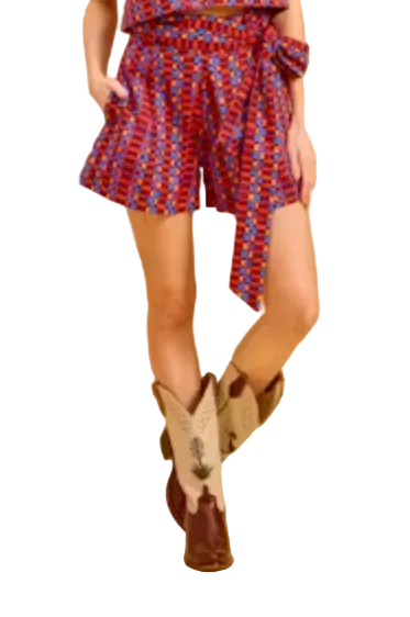 A woman modeling a sleeveless red and blue patterned romper with a matching tie belt, paired with brown and beige cowboy boots, against a solid beige studio background.