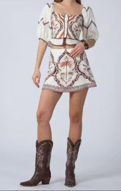 A woman modeling a two-piece bohemian-style outfit featuring a cream puff-sleeve crop top and matching mini skirt with brown and red paisley patterns, paired with dark brown cowboy boots, against a light gray studio background.