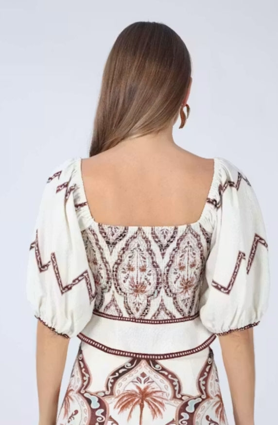 A woman viewed from the back wearing a cream-colored dress with brown and red bohemian-style embroidery and puff sleeves, set against a plain light gray background.