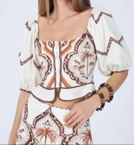 A woman modeling a cream-colored puff-sleeve crop top with brown and orange paisley embroidery, paired with a matching embroidered skirt, against a light gray studio background.