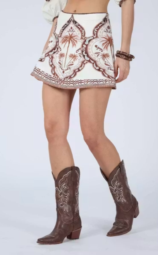 A model wearing a white western-style mini skirt with brown embroidered floral and geometric patterns, paired with brown leather cowboy boots, against a light gray studio background.